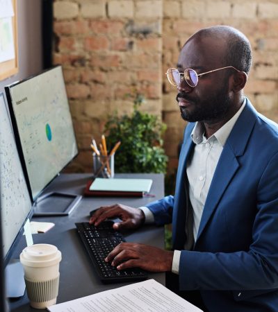 Manager working with charts on computer Serious African manager in eyeglasses sitting at his workplace at office and typing, he working with charts on computer